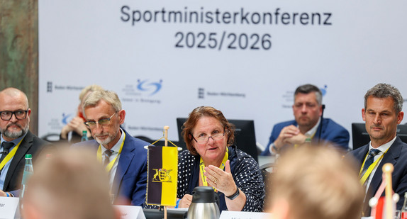 Mehrere Personen sitzen an langen Tischen in einer Halle. 
