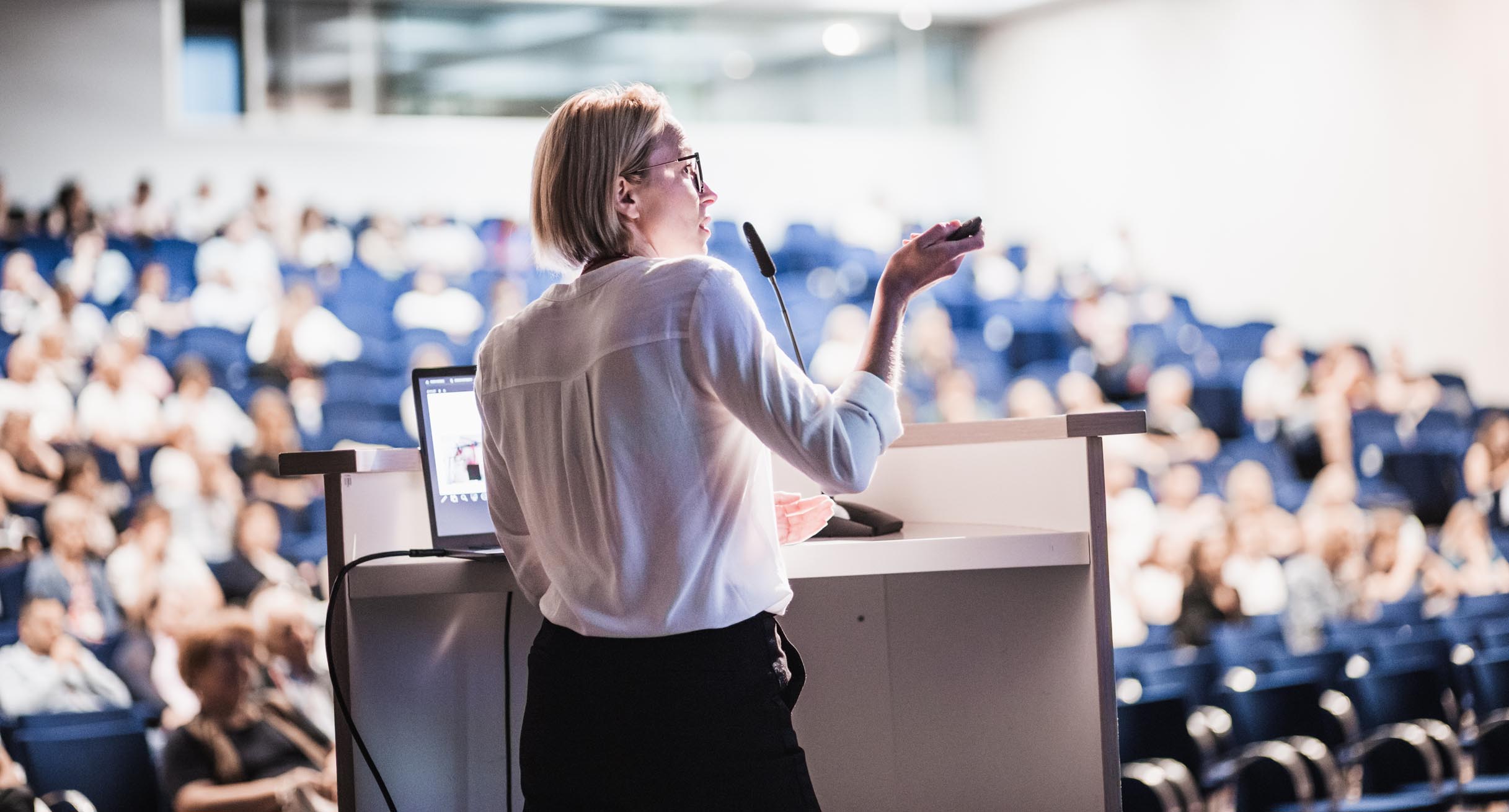 Female speaker giving a talk on corporate business conference. Unrecognizable people in audience at conference hall. Business and Entrepreneurship event.